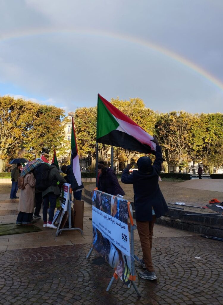 Photo du rassemblement avec un drapeau soudanais et en fond un arc-en-ciel.
